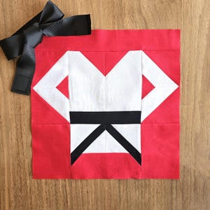 May include: A red and white fabric quilt block with a black belt design, representing a karate uniform.
