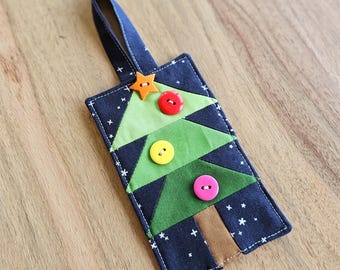 May include: A handmade Christmas tree ornament. The ornament features a patchwork design with a green tree, brown trunk, and colorful buttons. The background is dark blue with white star accents, and a blue loop for hanging.