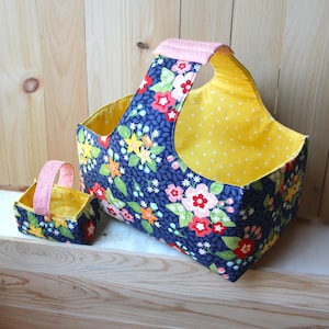 May include: Two fabric baskets, one large and one small, with a floral pattern on a navy blue background. The larger basket has a pink handle and yellow polka dot lining. The smaller basket has a pink handle and yellow polka dot lining.