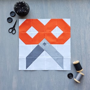 May include: A white fabric block with a geometric pattern in orange and grey. The block is made up of 16 smaller squares, with the orange squares forming an infinity symbol in the top row and the grey squares forming a chevron pattern in the bottom row.