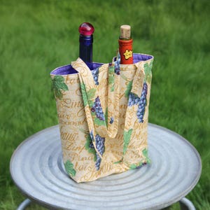 May include: A beige wine tote bag with a purple lining, holding two wine bottles. The bag has a grape and leaf pattern with script writing. One bottle has a red stopper, the other a dark blue bottle with a red stopper.