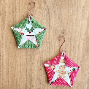 Puede incluir: Dos adornos navideños hechos a mano en forma de estrella. Una estrella es verde con un diseño de cupcake blanco y la otra es roja con un diseño de hombre de jengibre blanco.