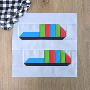May include: A square fabric quilt block featuring two pieced ship designs on a light gray background. Each ship has a black hull, a light blue bow, and a cabin section composed of vertical stripes in alternating green, red, and blue fabric.