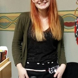 May include: A black and white patterned apron with multiple pockets, including a cell phone pocket. The apron is worn by a person with long red hair and glasses.