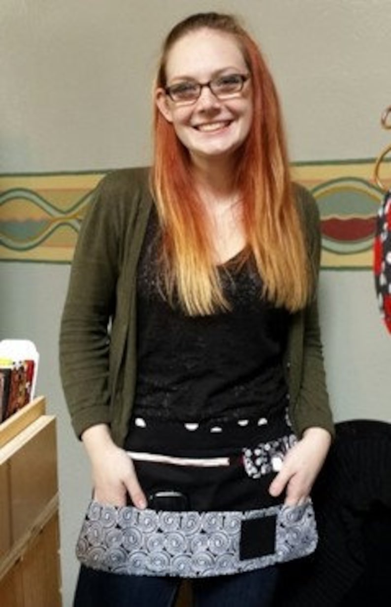 May include: A black and white patterned apron with multiple pockets, including a cell phone pocket. The apron is worn by a person with long red hair and glasses.