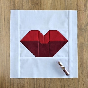 Puede incluir: Un bloque de tela con un diseño de labios rojos y burdeos sobre un fondo blanco. Un tubo de lápiz labial está al lado del bloque. El diseño de labios está hecho de formas geométricas y está centrado en la tela blanca.