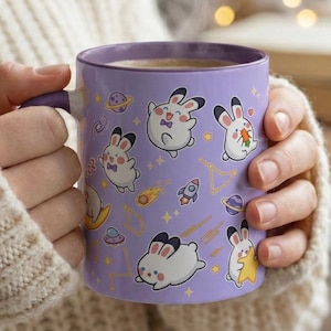 May include: A purple ceramic mug with a handle, featuring a repeating pattern of cartoon bunnies, planets, rockets, and stars. The mug has a purple interior and is held by a person wearing a cream-colored sweater.