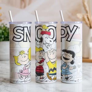 May include: Three white insulated tumblers with the word "SNOOPY" in black block letters. Each tumbler features a different illustration of Snoopy and other Peanuts characters. Each tumbler has a white straw.