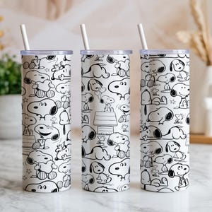 May include: Three white insulated tumblers with a black and white Snoopy cartoon character pattern. Each tumbler has a clear lid and a white straw. The design features Snoopy in various poses, including lying down and sitting.