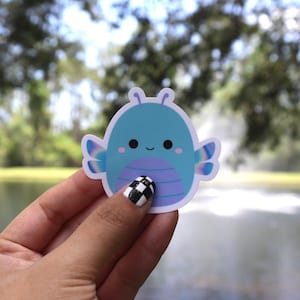 May include: A blue sticker featuring a cartoon-style insect with a smiling face. The insect has a light blue body, purple belly, and iridescent wings. The sticker is held in a hand with a black and white checkered nail design.