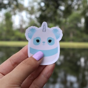 May include: A sticker featuring a panda-unicorn hybrid design. The sticker has a white base with a light blue stripe and accents. The panda-unicorn has black eyes, blue ears, and a silver horn. The sticker is held by a person.