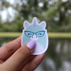 May include: A die-cut sticker featuring a cartoon unicorn with teal fur, a blue horn, and blue glasses. The unicorn has a white belly and is set against a white background. The sticker is held by a person.