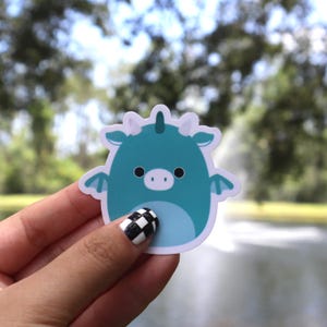 May include: A teal dragon sticker with a cute design. The sticker has a white outline, small wings, and a light blue belly. The background is blurred, showing a body of water and trees.