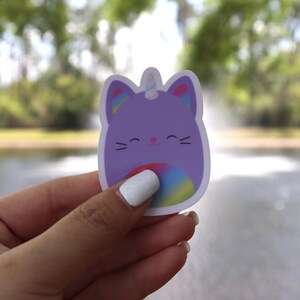 May include: A purple cat-unicorn sticker with a rainbow belly and accents. The sticker has a white border and a smiling face. The background is blurred, showing a body of water and trees.
