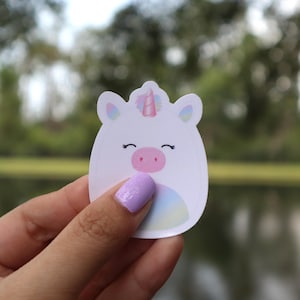 May include: A white unicorn sticker with a smiling face, pink nose, and a rainbow-colored belly. The unicorn has a pink and purple horn. The sticker is held by a hand with a light purple nail polish.