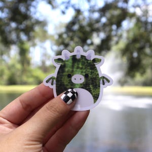 May include: A sticker featuring a green dragon-like creature with a white belly and small wings. The dragon has a textured pattern and a cute, round face. The sticker is held in a hand with a black and white checkered nail design.