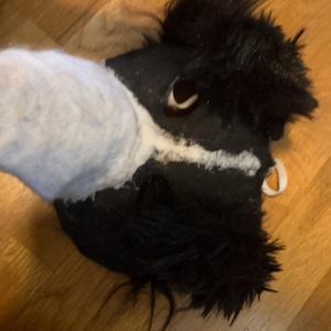 May include: A black and white animal mask featuring long, shaggy black faux fur around the ears and eyes, with a prominent white stripe extending from the snout to the forehead. The mask includes a dark nose and white elastic straps for securing.