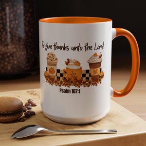 Może przedstawiać: Biały ceramiczny kubek z pomarańczowym wnętrzem i uchwytem. Kubek ma jesienny wzór z napojami kawowymi, dyniami oraz tekstem "I give thanks unto the Lord" i "Psalm 107:1".