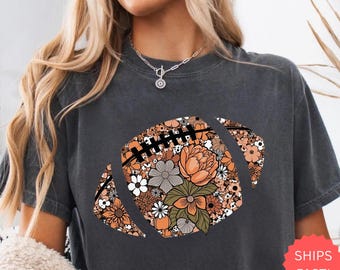 May include: Dark gray t-shirt featuring a floral football design in shades of orange, white, and green. The shirt has a crew neck and short sleeves. The word "PEPPER" is printed at the bottom of the shirt.