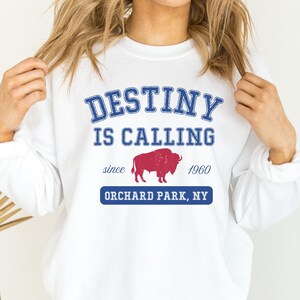 May include: White sweatshirt with the words 'DESTINY IS CALLING' in blue block letters. Below the text is a red buffalo graphic and the words 'since 1960'. Underneath is the text 'ORCHARD PARK, NY' in a blue rectangle.