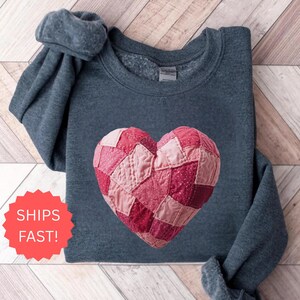 May include: A dark gray sweatshirt featuring a patchwork heart design in shades of pink and red. The heart is centrally placed on the chest. The sleeves are folded. The words "SHIPS FAST!" are in a red circle in the lower left corner.