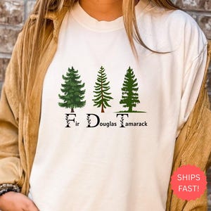 May include: Cream-colored t-shirt featuring three green fir trees with the text "Fir Douglas Tamarack" in a decorative font. The shirt has a relaxed fit and is made of a soft material.