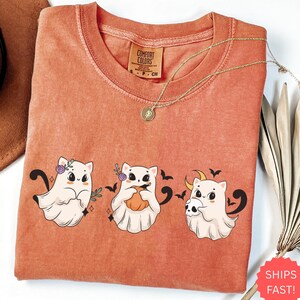May include: Orange Comfort Colors t-shirt featuring three cartoon ghost cats with Halloween-themed accessories. The cats are wearing floral crowns, holding pumpkins, and a skull. The shirt has a crew neck and a small tag that says "Comfort Colors".