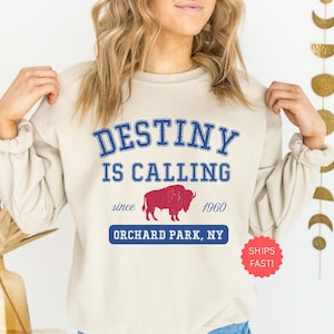 May include: A cream-colored sweatshirt with a blue and red graphic. The graphic reads 'DESTINY IS CALLING' above 'since 1960' and 'ORCHARD PARK, NY'. A red buffalo image is in the center. A red circle with 'SHIPS FAST!' is on the bottom right.