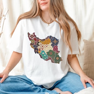 May include: White t-shirt featuring a colorful, patterned map of Iran. The map design incorporates various colors and patterns, including red, blue, yellow, and green. The shirt is paired with blue jeans.