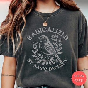 以下が含まれることがあります： 「RADICALIZED BY BASIC DECENCY」の文字と、花に囲まれた灰色の鳥のイラストがプリントされた濃いグレーのTシャツ。 ヴィンテージ感のあるウォッシュ加工が施されています。