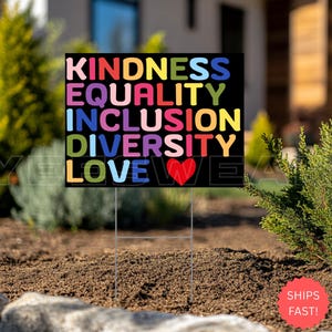 May include: A black yard sign with the words "KINDNESS, EQUALITY, INCLUSION, DIVERSITY, LOVE" in colorful block letters. A red heart replaces the letter "O" in "LOVE". The sign is displayed in a garden setting.