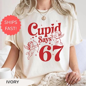 May include: Ivory-colored t-shirt with a red graphic that reads "Cupid Says 67" and features two cherubs with bows and arrows. The shirt has a crew neck and short sleeves. A red sticker that says "Ships Fast!" is visible.