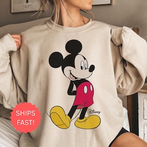 Op de afbeelding: Beige sweatshirt met een grote Mickey Mouse-afbeelding. Mickey wordt getoond in zijn klassieke pose, met zwarte oren, een wit gezicht, rode shorts, gele schoenen en witte handschoenen. Het Disney-logo staat rechtsonder.