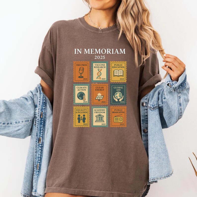 May include: Brown t-shirt with the text "IN MEMORIAM 2025" and postage stamp-style graphics. The stamps depict themes like free press, vaccine research, climate action, and public broadcasting. The shirt has rolled-up sleeves.