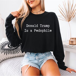 以下が含まれることがあります： 白字で「Donald Trump Is a Pedophile」と書かれた黒い長袖スウェットシャツ。スウェットシャツはデニムショーツと合わせて着用されています。画像は屋内撮影です。
