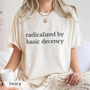 Puede incluir: Camiseta color marfil con el texto negro "radicalized by basic decency". La camiseta tiene mangas cortas y un corte holgado. Se sostiene una taza blanca en la mano. La palabra "Ivory" est&aacute; impresa en la parte inferior de la imagen.