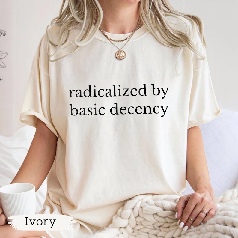 Puede incluir: Camiseta color marfil con el texto negro "radicalized by basic decency". La camiseta tiene mangas cortas y un corte holgado. Se sostiene una taza blanca en la mano. La palabra "Ivory" est&aacute; impresa en la parte inferior de la imagen.
