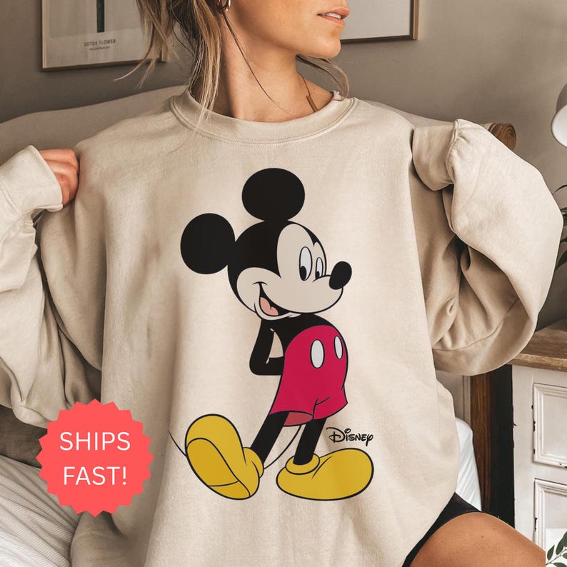 Op de afbeelding: Beige sweatshirt met een grote Mickey Mouse-afbeelding. Mickey wordt getoond in zijn klassieke pose, met zwarte oren, een wit gezicht, rode shorts, gele schoenen en witte handschoenen. Het Disney-logo staat rechtsonder.