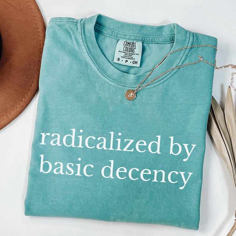 Puede incluir: Una camiseta Comfort Colors turquesa con la frase "radicalized by basic decency" en blanco. Un collar dorado con un peque&ntilde;o colgante descansa sobre la camiseta. Un sombrero marr&oacute;n y hojas secas est&aacute;n en el fondo.