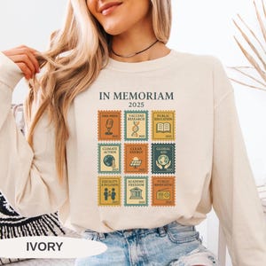 May include: Ivory-colored sweatshirt with a graphic of postage stamps featuring icons and text including "IN MEMORIAM 2025", "Free Press", "Vaccine Research", and "Public Education". The word "IVORY" is printed at the bottom.