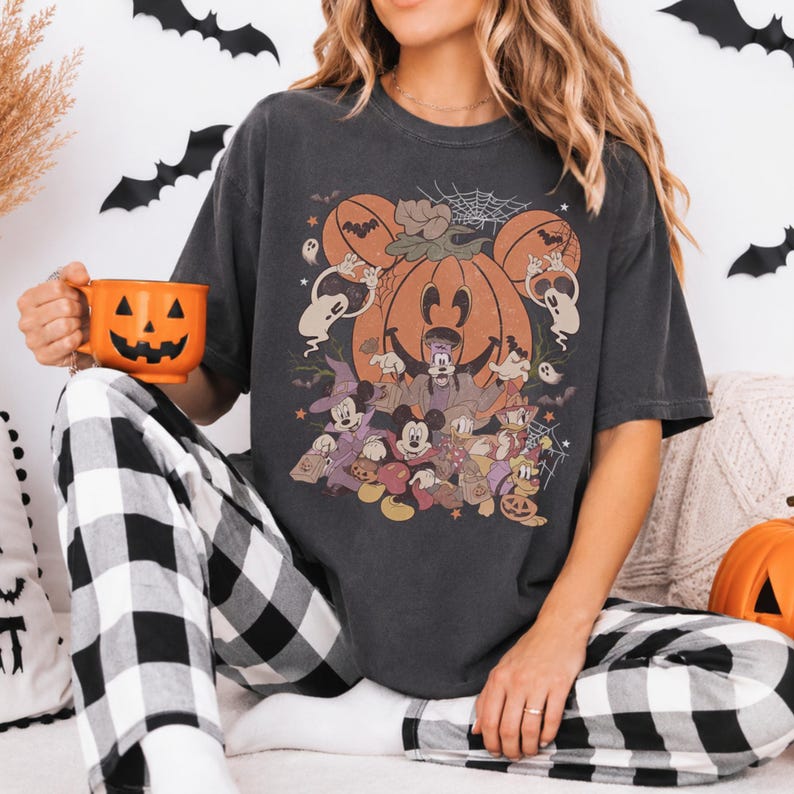 May include: A charcoal gray short-sleeve t-shirt featuring a large graphic print of various cartoon characters dressed in Halloween costumes, including a central pumpkin, ghosts, and bats. The shirt is paired with black and white plaid pants.