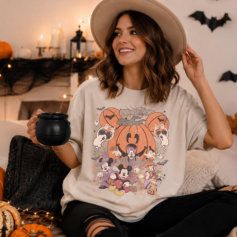 May include: A beige short-sleeve t-shirt featuring a graphic print of Disney characters in Halloween costumes, including Mickey Mouse, Minnie Mouse, and Goofy, arranged around a large orange pumpkin with Mickey-shaped ears and small ghosts.