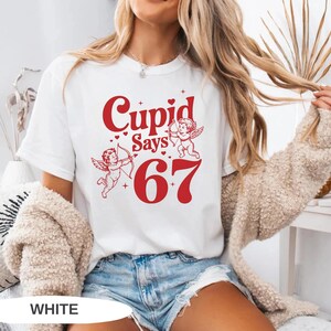 May include: White t-shirt with a red graphic that says "Cupid Says 67" with two cherubs and hearts. The shirt is a casual style, suitable for everyday wear.