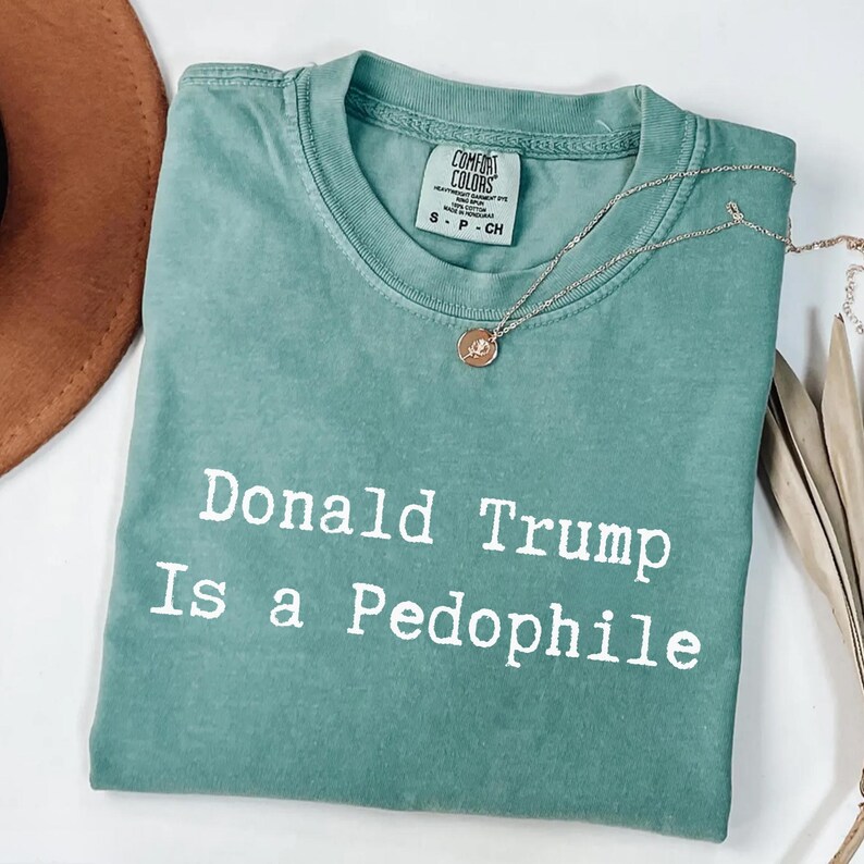 以下が含まれることがあります： 「Donald Trump Is a Pedophile」の白文字がプリントされた、シーフォームグリーンのTシャツ。Tシャツは折りたたまれており、Comfort Colorsのタグが付いています。ペンダント付きのシルバーネックレスと茶色の帽子も写っています。