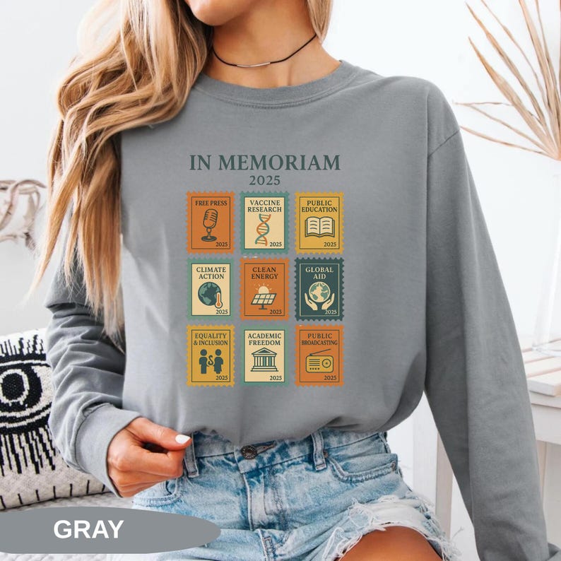May include: Gray long-sleeve sweatshirt with the text "IN MEMORIAM 2025" and postage stamp-style graphics. The graphics include images related to free press, vaccine research, public education, climate action, clean energy, global aid, equality & inclusion, academic freedom, and public broadcasting.