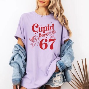 May include: A light purple t-shirt with the text "Cupid Says 67" in red. The design includes two cherubs with bows and arrows. The shirt is a casual style.