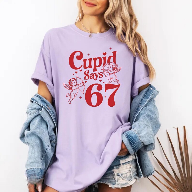 May include: A light purple t-shirt with the text "Cupid Says 67" in red. The design includes two cherubs with bows and arrows. The shirt is a casual style.