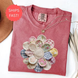 May include: A dusty rose Comfort Colors t-shirt featuring a large, appliquéd flower design. The flower is made from various fabric scraps in floral, gingham, and lace patterns. A silver necklace with a small pendant rests on the shirt. The shirt has a tag that says S-P-CH.