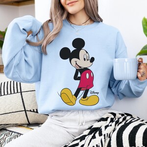 Op de afbeelding: Lichtblauw sweatshirt met een Mickey Mouse-afbeelding. De tekenfilmfiguur is afgebeeld in zijn klassieke outfit: rode shorts, gele schoenen en zwarte handschoenen. Het Disney-logo staat eronder.