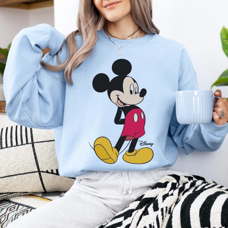 Op de afbeelding: Lichtblauw sweatshirt met een Mickey Mouse-afbeelding. De tekenfilmfiguur is afgebeeld in zijn klassieke outfit: rode shorts, gele schoenen en zwarte handschoenen. Het Disney-logo staat eronder.